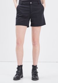 Mustat puuvillashortsit, joissa on keskikorkea vyötärö, etutaskut ja yksi hopea nappi. Yhdistetty mustien nilkkureiden kanssa, joissa on solkiyksityiskohtia.