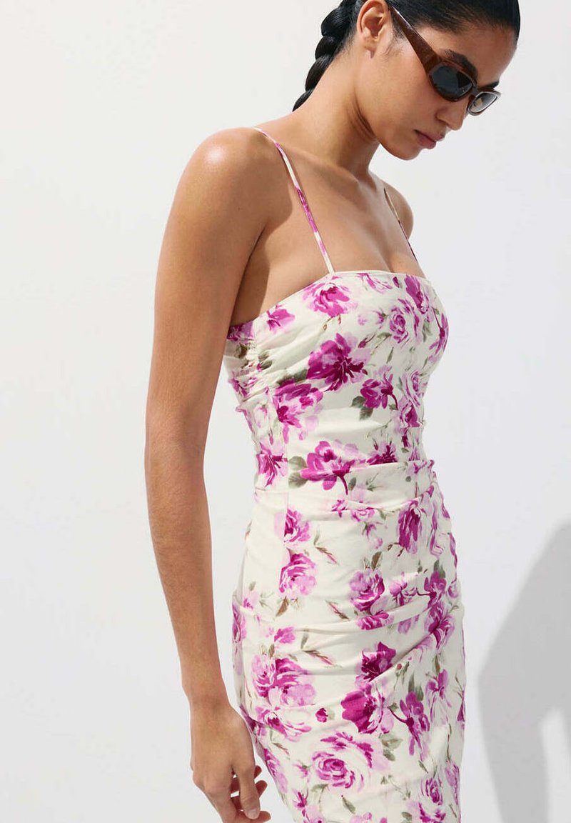 Femme aux cheveux tressés porte des lunettes de soleil et une robe blanche près du corps à motif floral rose, debout devant un fond uni clair.