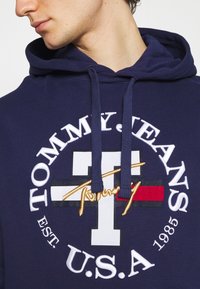 Homme portant un hoodie bleu marine avec "TOMMY JEANS U.S.A. EST. 1985" et un grand logo T avec signature et des bandes rouges, blanches et noires sur la poitrine.