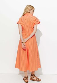 Robe rayée orange et blanche avec des manches courtes, un col et un ourlet évasé. Portée avec des sandales brunes à motifs. Texture de tissu lisse.