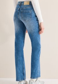 Personne portant un jean en denim bleu taille haute avec poches arrière, associée à un haut blanc à manches longues et des baskets argentées, debout de profil.