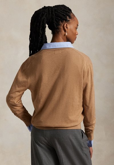 Femme aux cheveux tressés en queue de cheval, portant un pull camel par-dessus une chemise bleue à col et un pantalon gris, dos tourné sur un fond uni.