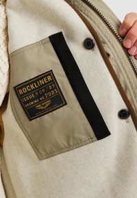 Beige yttertyg med en mjuk struktur; svart och olivgrön lapp med gul text; brun dragkedjekant; synligt knappdetalj.