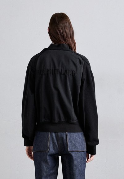 Fekete bomber dzseki vastag kötött gallérral és mandzsettákkal, a hátán "HELMUT LANG" hímzéssel. Kék farmernadrággal párosítva.