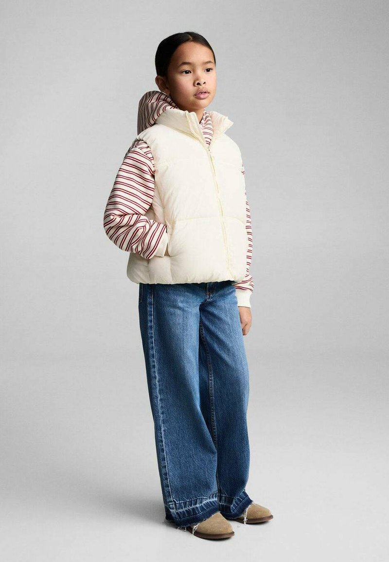 Puffige cremefarbene Weste über einem rot-weiß gestreiften Langarmshirt, kombiniert mit locker sitzenden blauen Jeans und beigen Schuhen. Einfaches Design.