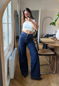 Blugi din denim albastru închis, cu talie înaltă și croială cu picioare largi, evidențiați de o textură fină a țesăturii și o talie ajustată.