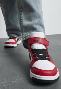 Baskets rouges et blanches avec des accents noirs, en matériaux cuir et synthétiques, avec un toe perforé et une sangle ajustable. Laces et logo visibles.