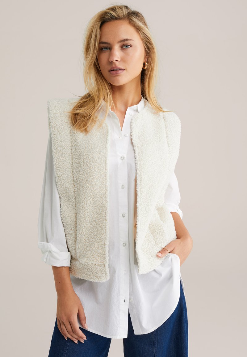 Fleece jopice v bež barvi z mehkobnim, strukturiranim zaključkom in odprtim sprednjim delom, nameščene čez svež belo srajco z gumbi. Priložnostni kroj.