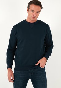 Sötétkék crewneck pulóver hosszú ujjakkal, bordás mandzsettákkal és texturált felülettel. Kis háromszög alakú díszítés található a nyaknál.