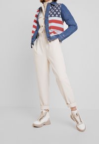 Chaqueta de mezclilla con rayas rojas y blancas, diseño de bandera americana en la parte delantera, combinada con pantalones taper de color crema y zapatillas beige.