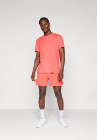 Coral activewear set featuring a T-shirt and shorts. T-shirt has a crew neck; shorts have a logo on the side. Worn with white sneakers.