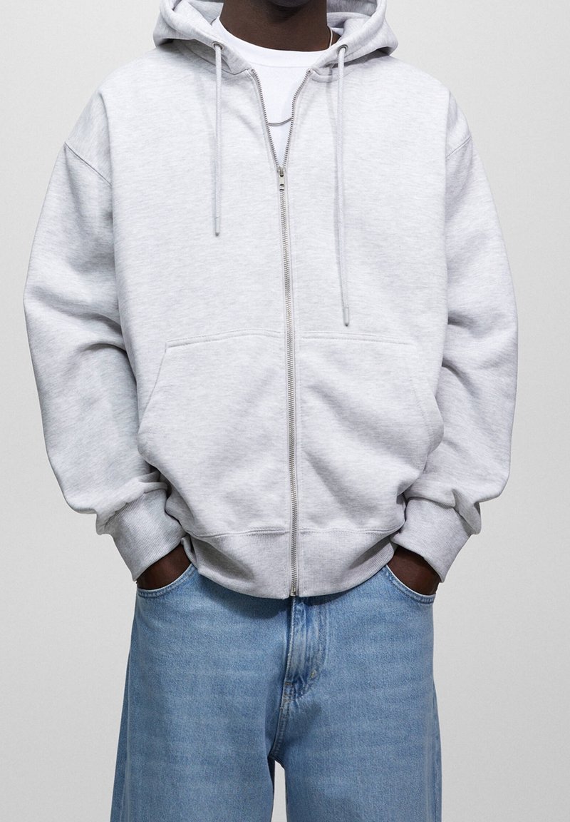 Personne portant un sweat à capuche gris clair zippé avec des cordons, une chemise blanche en dessous et un jean bleu, les mains dans les poches, debout devant un fond uni.