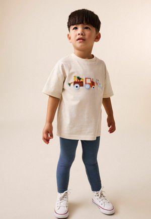 Ung dreng iført en cremefarvet t-shirt med et farverigt motiv af en traktor og en ko, blåstribede leggings og hvide sneakers, der står mod en ensfarvet baggrund.