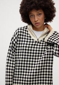Maglione lavorato a maglia con un motivo geometrico in bianco e nero, dotato di colletto con mezza zip e polsini a coste. Materiale misto lana dalla texture interessante.
