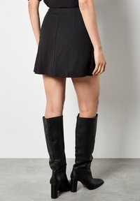 Schwarzer Rock mit ausgestelltem Saum, aus einem glatten Material gefertigt. Kombiniert mit knielangen schwarzen Lederstiefeln mit Blockabsatz.