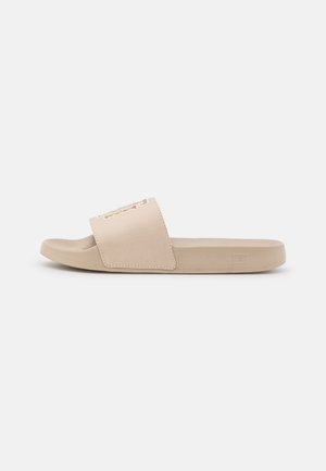 Beige slide-sandaal met brede band en platte zool, getoond in profiel op een neutrale achtergrond.