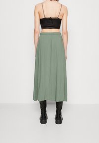 Femme se tenant de dos, portant un crop top noir, une jupe midi vert clair et des bottes montantes noires.