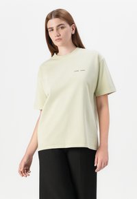A young woman poses confidently, wearing a light green oversized t-shirt with "SAMSØE" printed on the front and black pants.