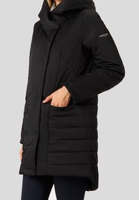 Žena v černém prošívaném zimním kabátu s vysokým límcem, bočními kapsami a logem Finn Flare na horním rukávu.