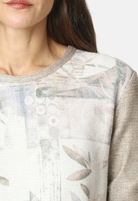 Mujer vistiendo un suéter texturizado y estampado con diseños geométricos y florales en tonos beige, blanco y pasteles suaves, mostrado desde los hombros hasta la barbilla.