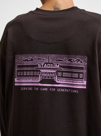 Osoba nosi crnu majicu s ružičastom grafikom stadiona i tekstom "SERVING THE GAME FOR GENERATIONS" na leđima.