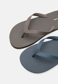 Δύο υφασμένα λαστιχένια σαγιονάρες με δέστρες για τα δάχτυλα, μία καφέ και μία μπλε, τοποθετημένες σε λευκό φόντο.