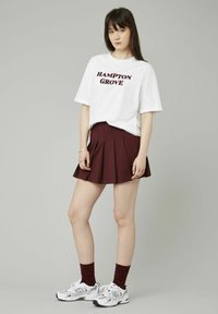 T-shirt en coton blanc avec "HAMPTON GROVE" imprimé en bordeaux foncé. Jupe plissée bordeaux, associée à des baskets blanches et noires et des chaussettes bordeaux.