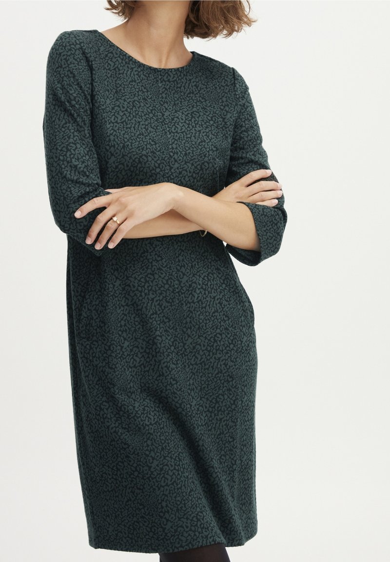 Femme aux cheveux courts portant une robe verte foncée à motifs, longueur genou, manches trois-quarts, bras croisés devant un fond blanc.