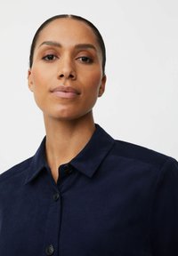 Camicia button-up blu navy con colletto, tessuto testurizzato e sottile lucentezza. Presenta grandi bottoni e una vestibilità strutturata.