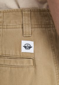 Khakihosen mit glatter Textur und standardmäßiger Passform. Enthält ein sichtbares weißes Etikett mit dem "Dockers"-Logo und einer einzelnen Gesäßtasche.