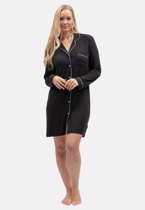 Femme aux longs cheveux blonds portant une chemise de nuit noire boutonnée avec un liseré blanc, debout pieds nus sur un fond clair uni.