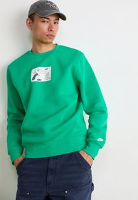 Felpa verde con un'applicazione grafica che presenta illustrazioni, polsini a coste e il logo Nike sulla manica. Abbinata a pantaloni cargo scuri.
