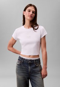 Valge lühendatud t-särk lühikeste varrukatega, mille keskel on peen Calvin Kleini logo, kombineeritud kõrge vöökohtadega tuhmide siniste teksadega.