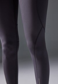 Zwarte atletische leggings met een gladde textuur, voorzien van contrasterende stiksels voor vormdefinitie en een reflecterende accent nabij de enkel.