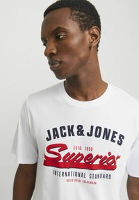 White cotton t-shirt featuring bold navy and red text, including "JACK & JONES SUPERIOR," and a round neckline.