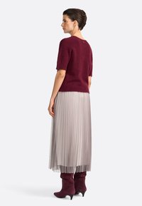 Maglione corto a maniche corte color bordeaux, gonna plissettata grigia chiara e stivali alti fino al ginocchio in suede bordeaux con punta affilata, visti da dietro.