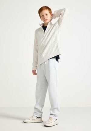 Punatukkainen poika, joka pukeutunut valkoiseen letitettyyn villapaitaan, tummansiniseen paitaan, vaaleanharmaisiin collegehousuihin ja kermaan sävyisiin lenkkareihin seisoo käsi päänsä takana.