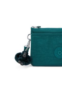 Teal stofpose med en tekstureret overflade, der har en lynlås lukning og en sort, fluffy gorilla charm hæftet på. Emblem på forsiden.