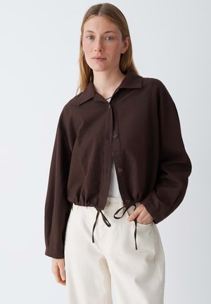 Frau mit langen blonden Haaren trägt eine dunkelbraune Jacke mit Knöpfen und Kordelzug sowie weiße, hoch taillierte Hose vor einem schlichten Hintergrund.