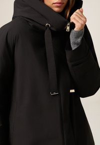 Zwart pufferjack met een hoge kraag, voorzien van een rits en een koordsluiting. Gladde textuur met gouden hardware accenten.