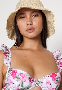 Biquíni floral com alças franzidas, apresentando uma base branca decorada com flores rosas e verdes. Acompanhado de um chapéu de palha natural em formato de balde.