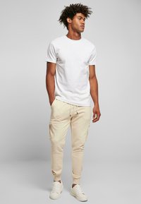 Tricou alb cu guler rotund, asortat cu pantaloni cargo bej cu buzunare laterale, completat cu adidași albi, pe un fundal gri simplu.