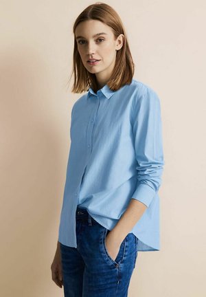 Light blue long-sleeve shirt with a button-up collar, featuring a relaxed fit and textured fabric, paired with dark denim jeans.
