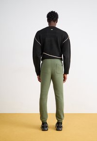 Sort sweater med cremefarvet syning, parret med olivengrønne sweatpants med lomme og tapered manchetter, sat mod en neutral baggrund.