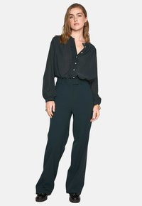 Maglietta nera a maniche lunghe con dettagli in bottone e texture sottile, abbinata a pantaloni high-waisted verde scuro e scarpe nere.