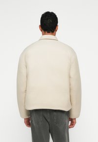 Šviesiai ruda pūkinė striukė su apykakle, plačiomis rankovėmis ir lygiu paviršiumi. Dėvima ant pilkų džinsų, rodo minimalistinį dizainą.