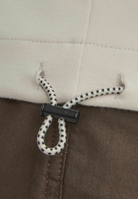 Un cordon de strângere elastic cu un toggle din plastic negru pe un material bej deschis, contrastând cu un material maro închis de dedesubt.