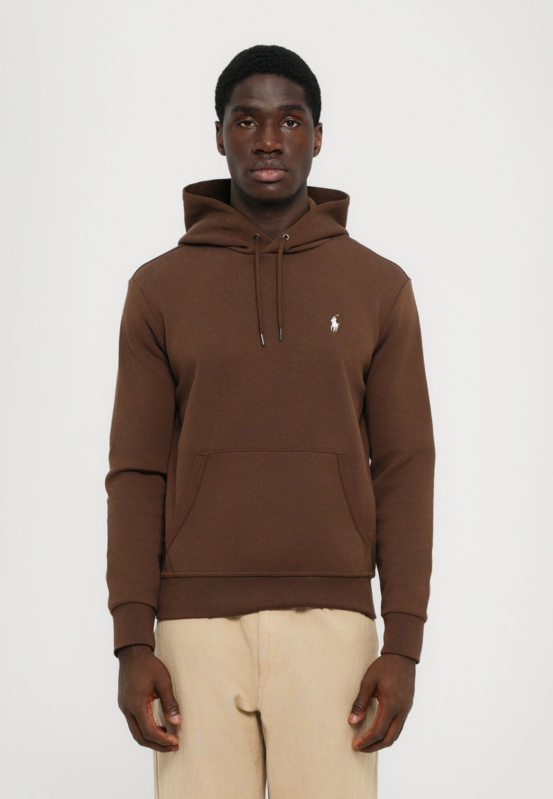 Hoodie castanho feito de um tecido macio, com um bolso frontal, capuz ajustável por cordão e um pequeno logotipo branco no peito. Combinado com calças beje claras.