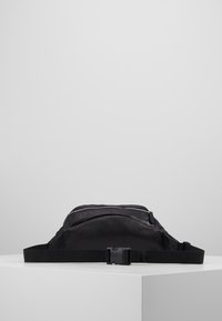 Musta nailoninen vyölaukku, jossa on tekstuuripinta, vetoketjusuljenta ja säädettävä hihna. Kompakti, suorakulmainen muotoilu soveltuu matkustamiseen.