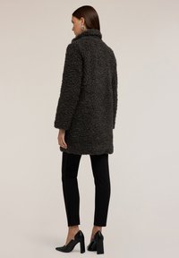 Uhlově šedé nadýchané svrchní oblečení s vysokým límcem a dlouhými rukávy, doplněné černými úzkými kalhotami a černými špičatými lodičkami.